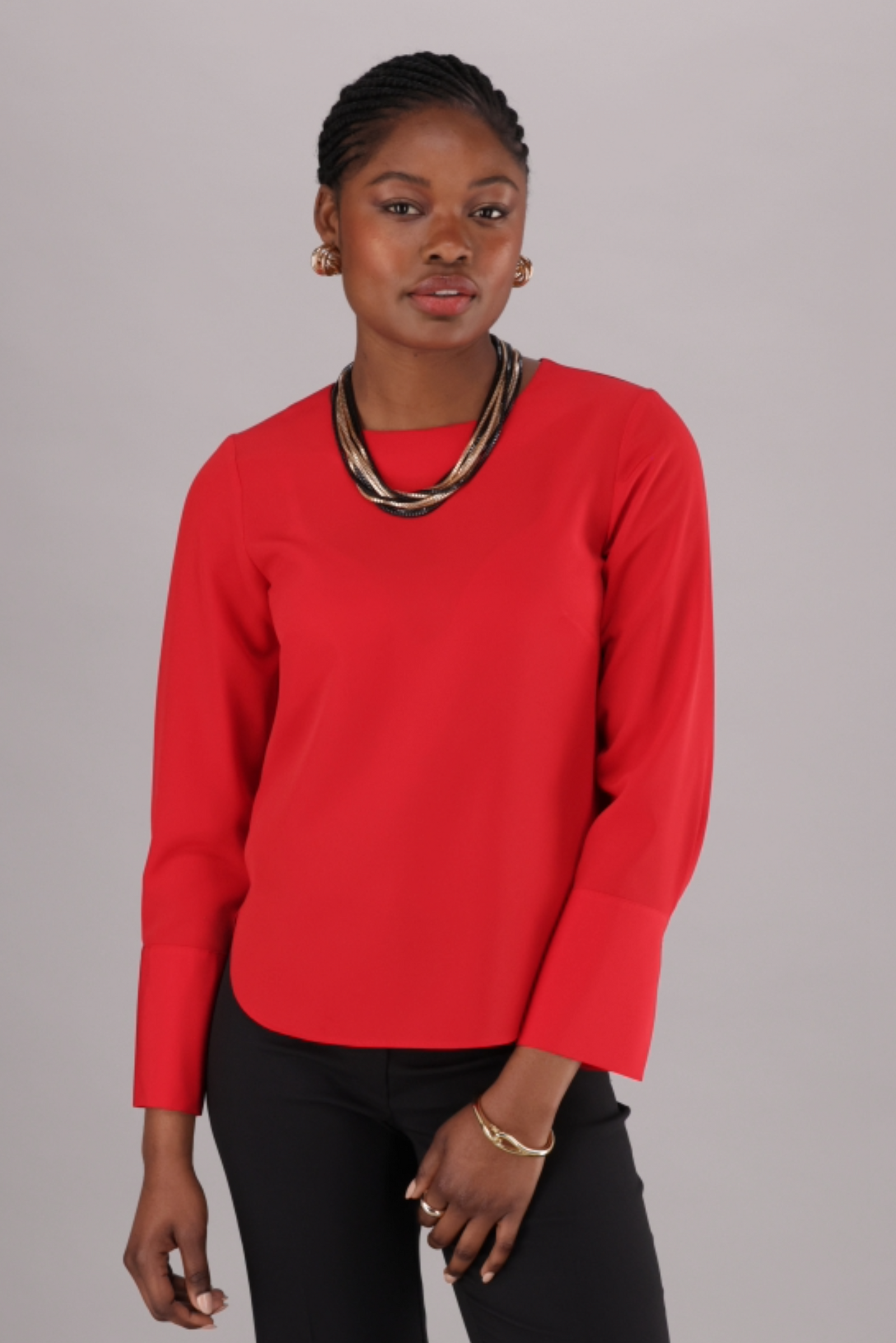 A woman wearing a red, long-sleeved blouse with a round neckline and scalloped hem, paired with black pants. Accessories include a patterned neck scarf and earrings.